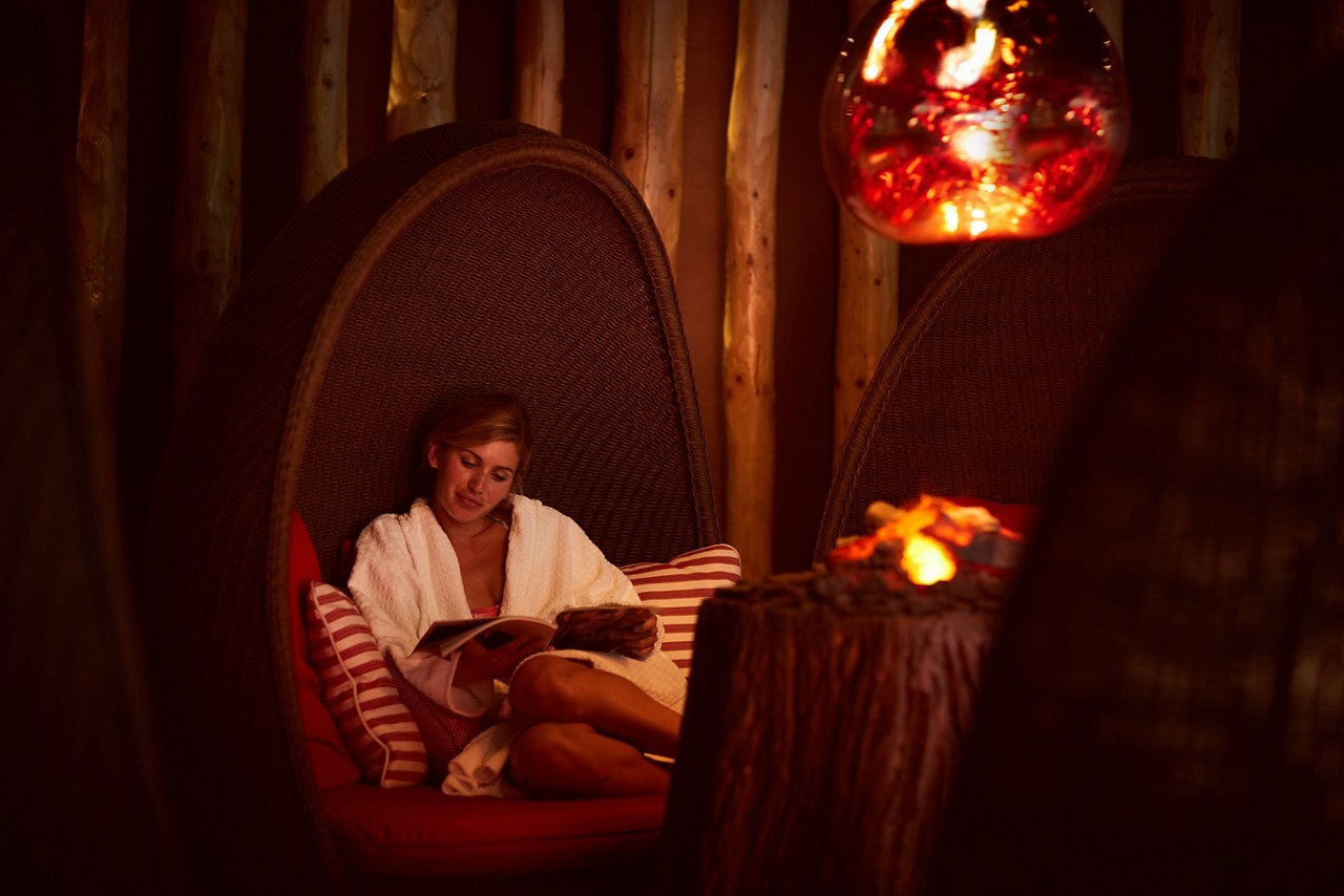 Woman snuggled up as she reads by a glowing fire inside the Fireside Relax.